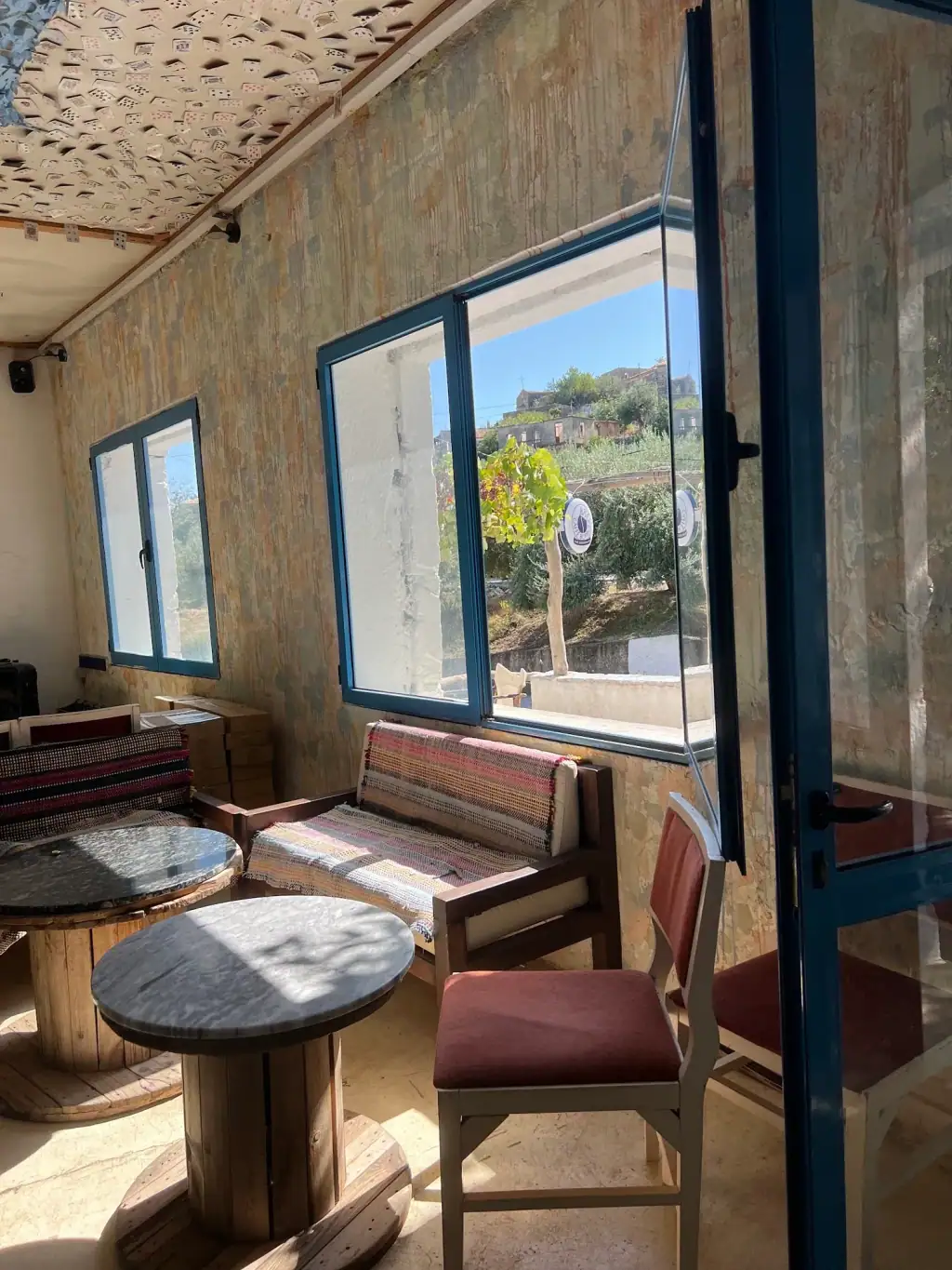 Himarë old town on its hill, seen through a blue-framed window at Bar 300, with a striped bench and marble tables inside
