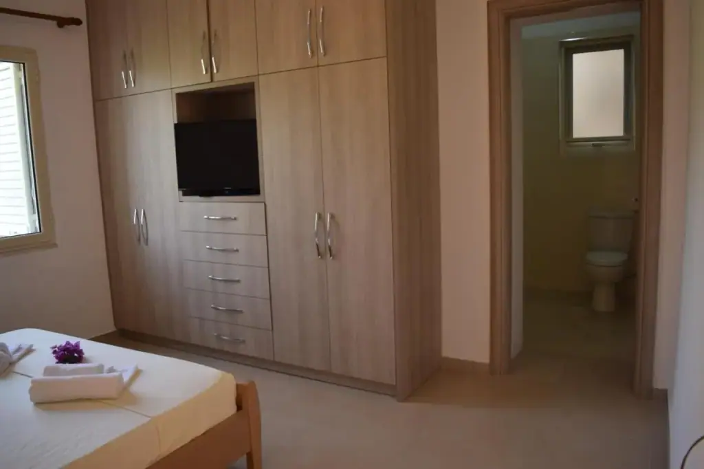 A fitted wall of pale wood wardrobes and drawers with a television recessed into it, a door to the bathroom beside
