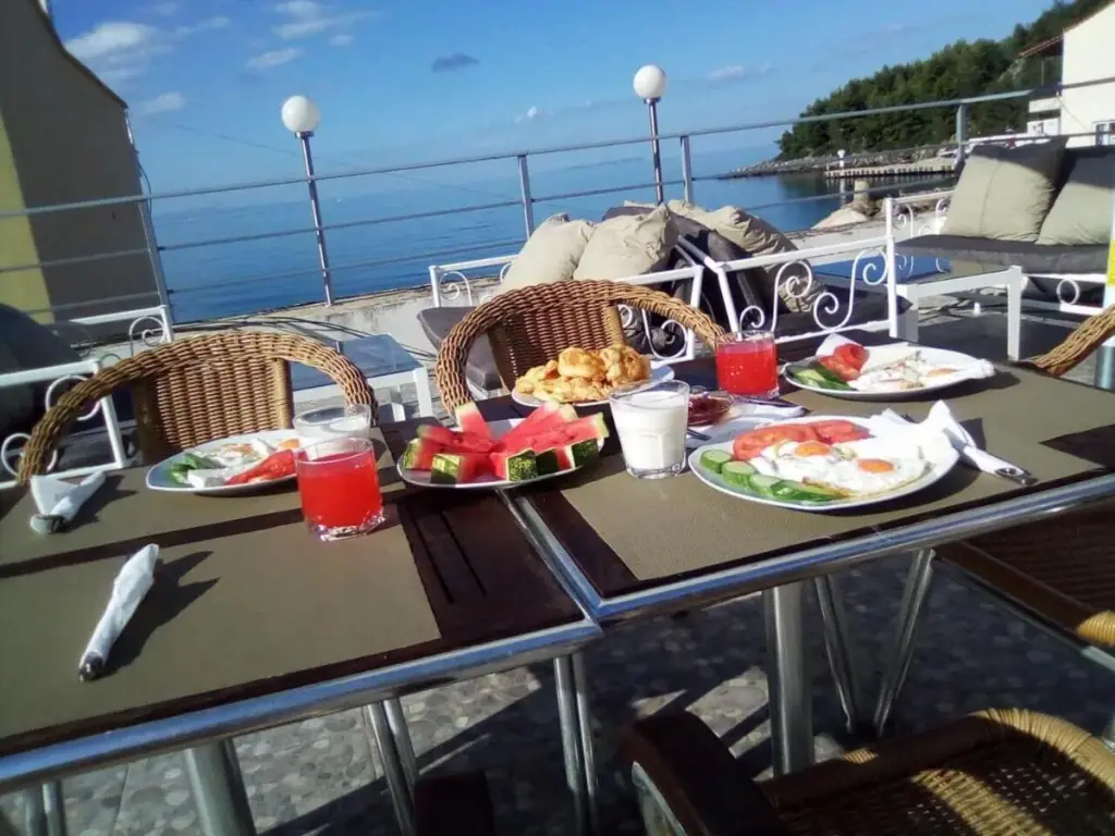 Breakfast laid on the roof terrace in morning sun — fried eggs, tomato, cucumber, white cheese, watermelon and juice, with the bay behind.