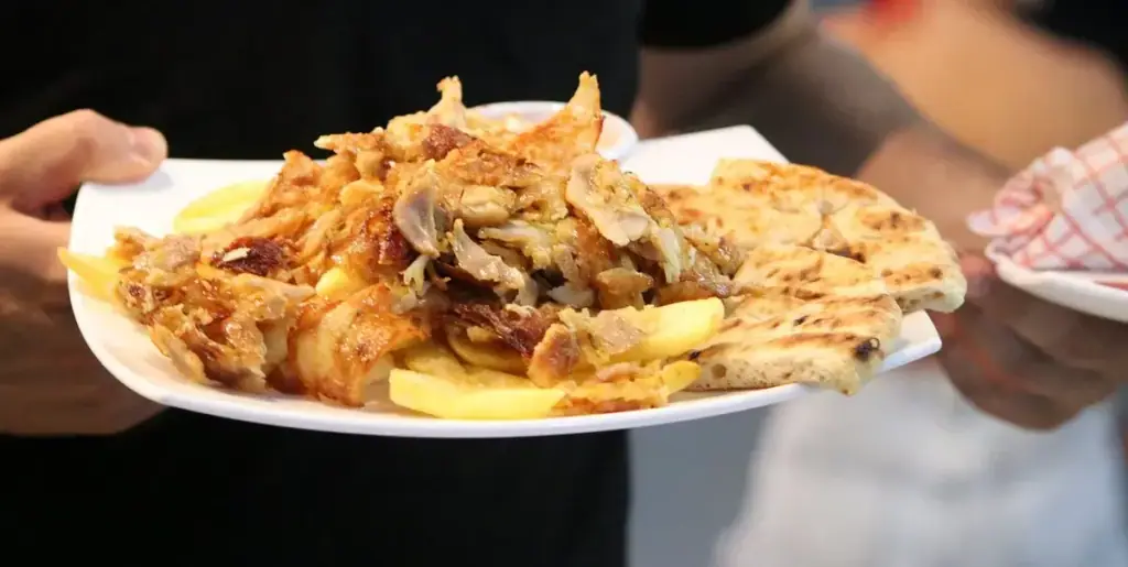 A white plate of carved meat over chips with grilled flatbread and a small pot of sauce, held in a hand, with a second plate lined with red-checked paper behind it