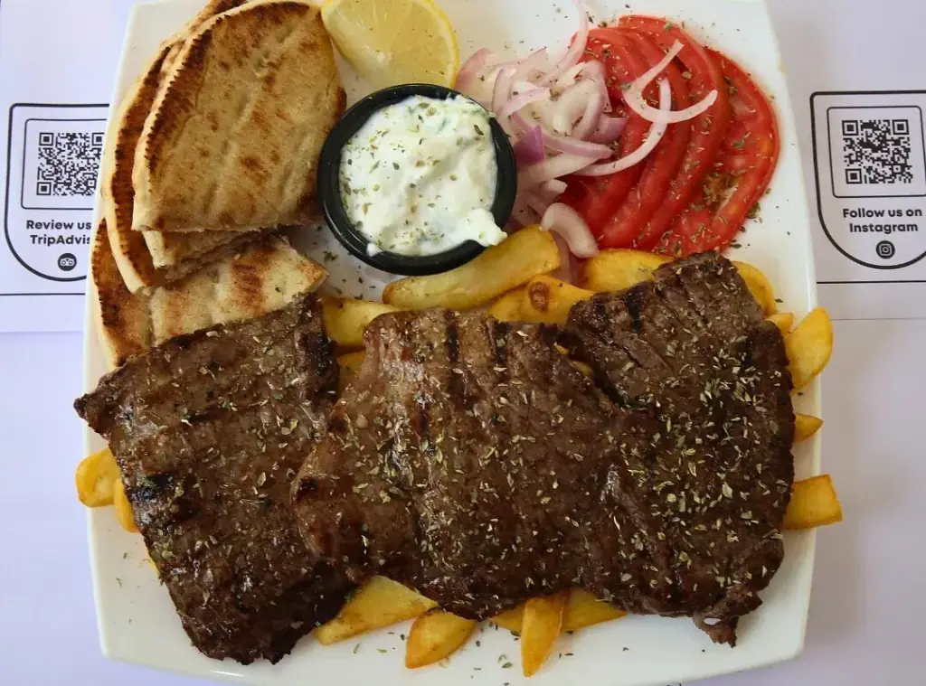 A white plate of three grilled cuts of meat over chips with grilled flatbread, a lemon wedge, sliced tomato, sliced red onion and a pot of white sauce, on a placemat printed with the venue's TripAdvisor and Instagram QR codes
