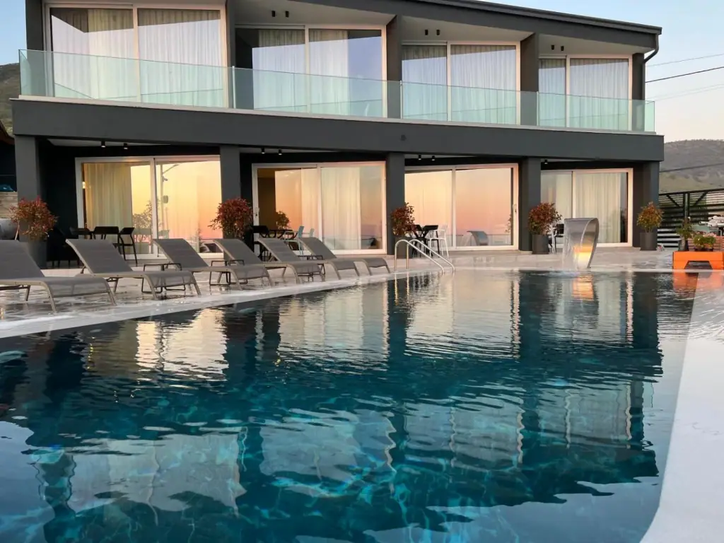 The graphite front of Casa Del Sol at dusk, its lit rooms and a peach sky doubled in the still pool, with grey sun loungers along the coping and a chrome water blade spilling in at the right