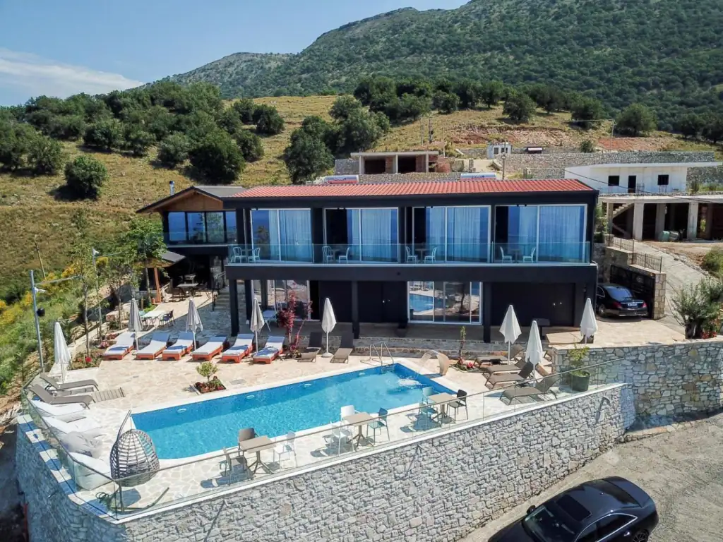 The whole property seen from above: a dark two-storey block under a terracotta pantile roof, its pool and flagstone terrace carried on a stone-clad retaining wall cut into a hillside of olive trees and dry grass