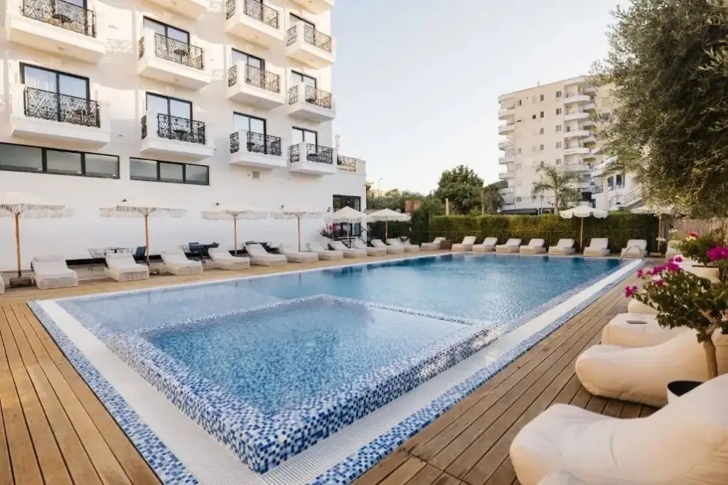 The outdoor pool at Chimaera Hotel, mosaic-tiled to the waterline, with a timber deck, white loungers and fringed parasols along the hotel wall