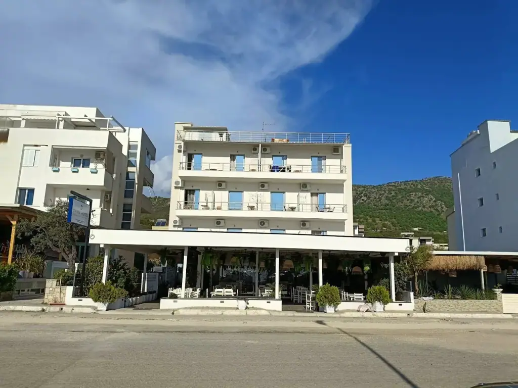 The four-storey white Dhima Hotel, three floors of balconies over a restaurant terrace under a flat canopy, and a green ridge behind