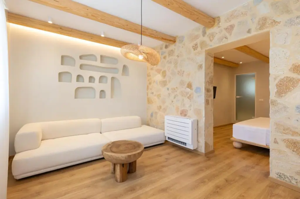 A sitting area in a room at Dimitri Hotel: a long cream sofa under a wall of moulded plaster niches, a wicker pendant, pine ceiling beams and a honey limestone wall