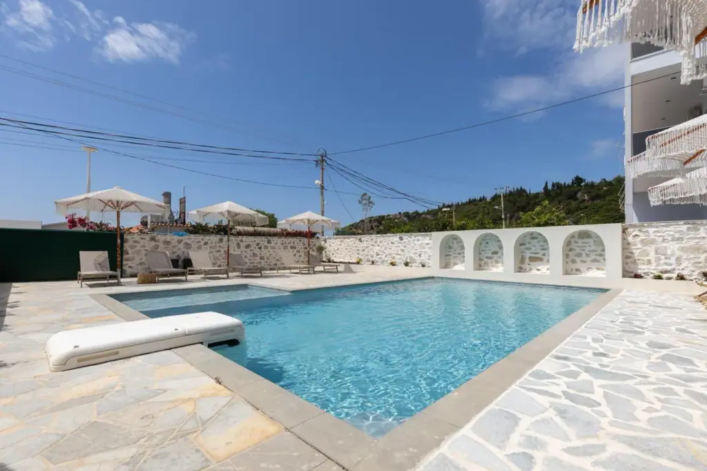 The outdoor pool at Dimitri Hotel on its crazy-paved terrace, loungers and fringed parasols along a drystone wall and the wooded ridge behind