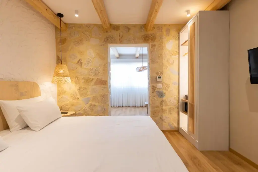 A bed at Dimitri Hotel against a wall of honey-coloured limestone rubble under pine beams, with a fringed cane pendant beside the pillows