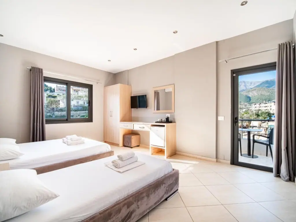 A room with two single beds, a desk and a balcony looking towards the mountains behind Himarë