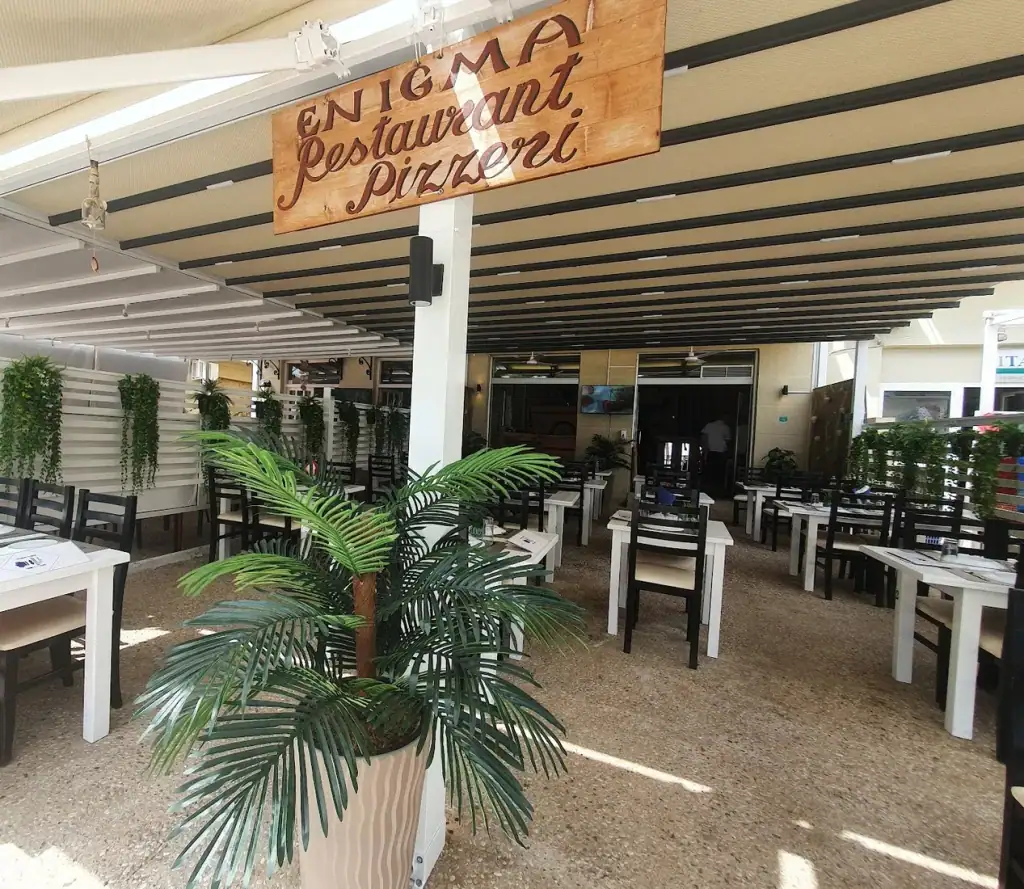 The covered terrace at Enigma: a cream retractable awning on dark battens, white tables and black slatted chairs, white slat screens hung with trailing greenery, and the painted board overhead