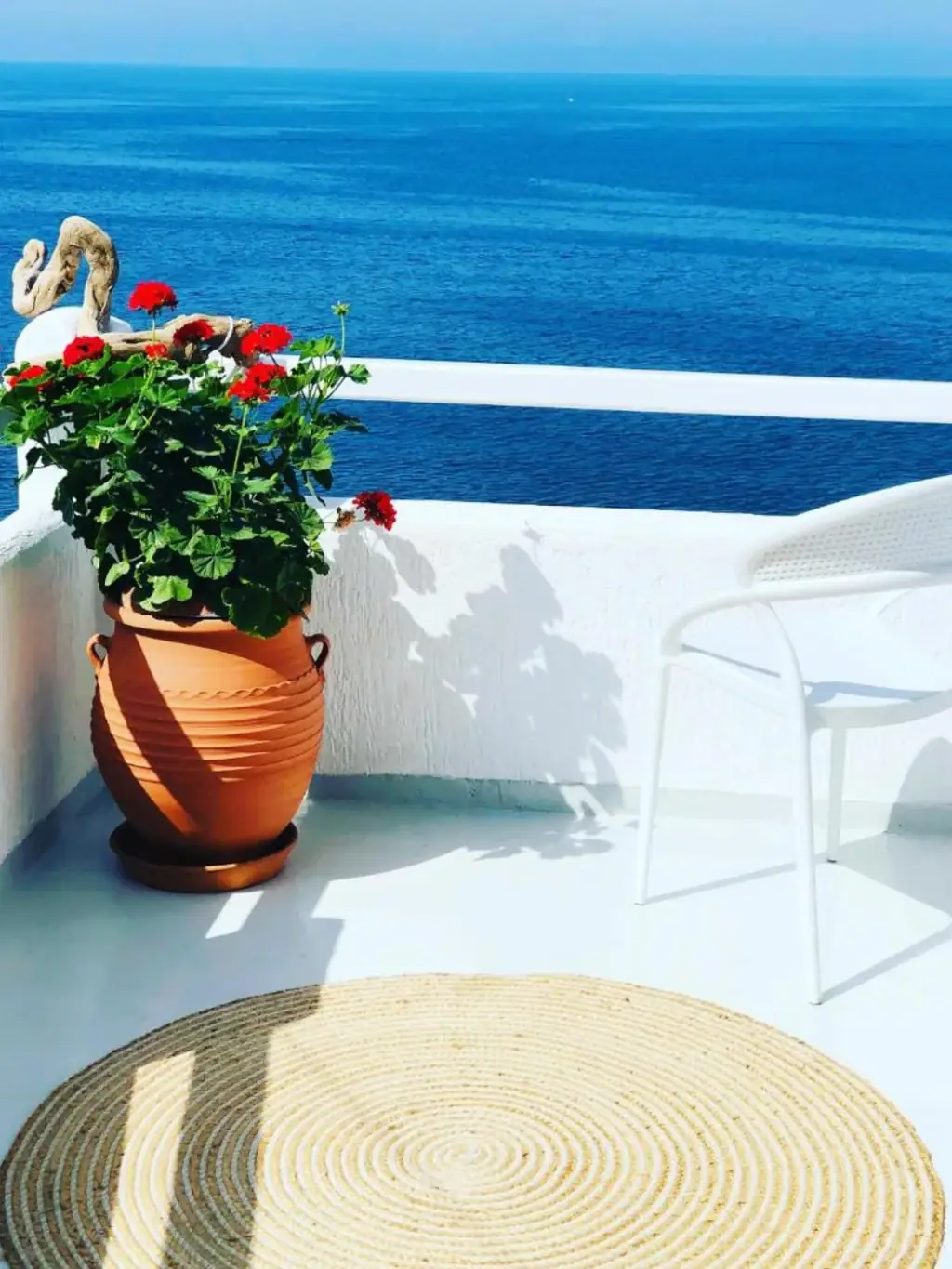 Red geraniums in a terracotta pot throwing a hard shadow across a white balcony wall above deep blue sea, with a round jute rug