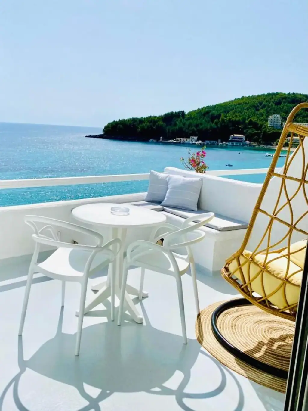 A white table and chairs, a cushioned bench and a hanging rattan chair on a balcony above the bay at Geo & Art Boutique Hotel