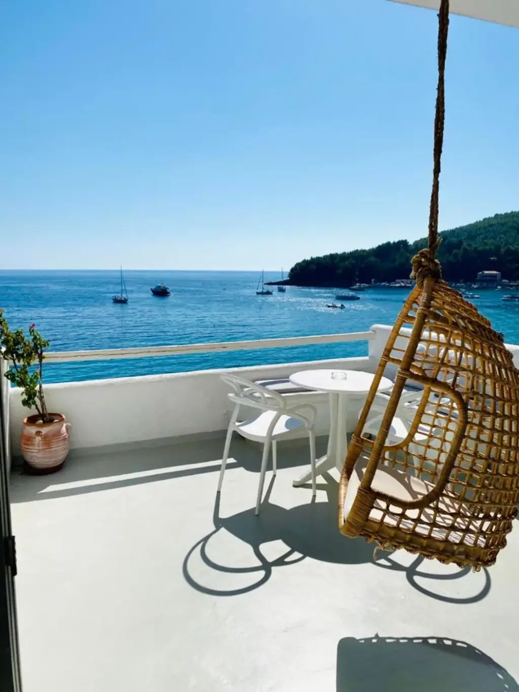 A hanging rattan chair, a white café table and a terracotta pot on a balcony looking out to moored boats and a wooded headland