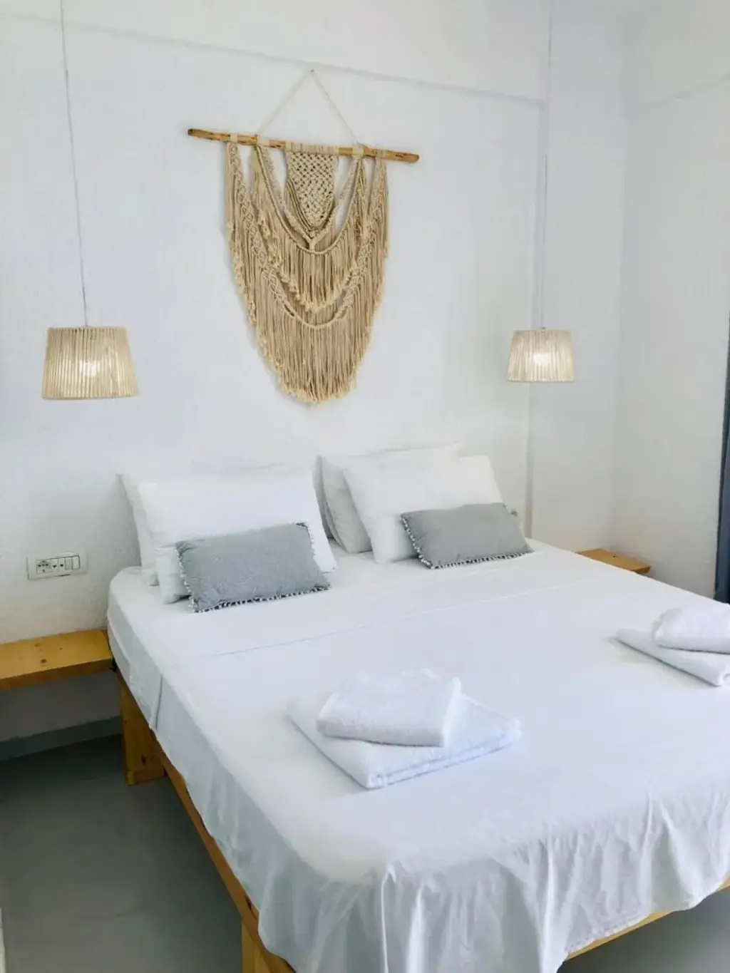 A pine bed in white linen beneath a macramé wall hanging, flanked by two woven pendant lamps