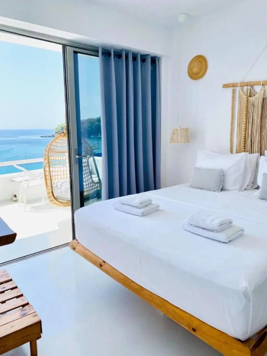 A pine-framed bed with white linen beside an open sliding door onto a balcony, a rattan pendant lamp and a woven wall hanging