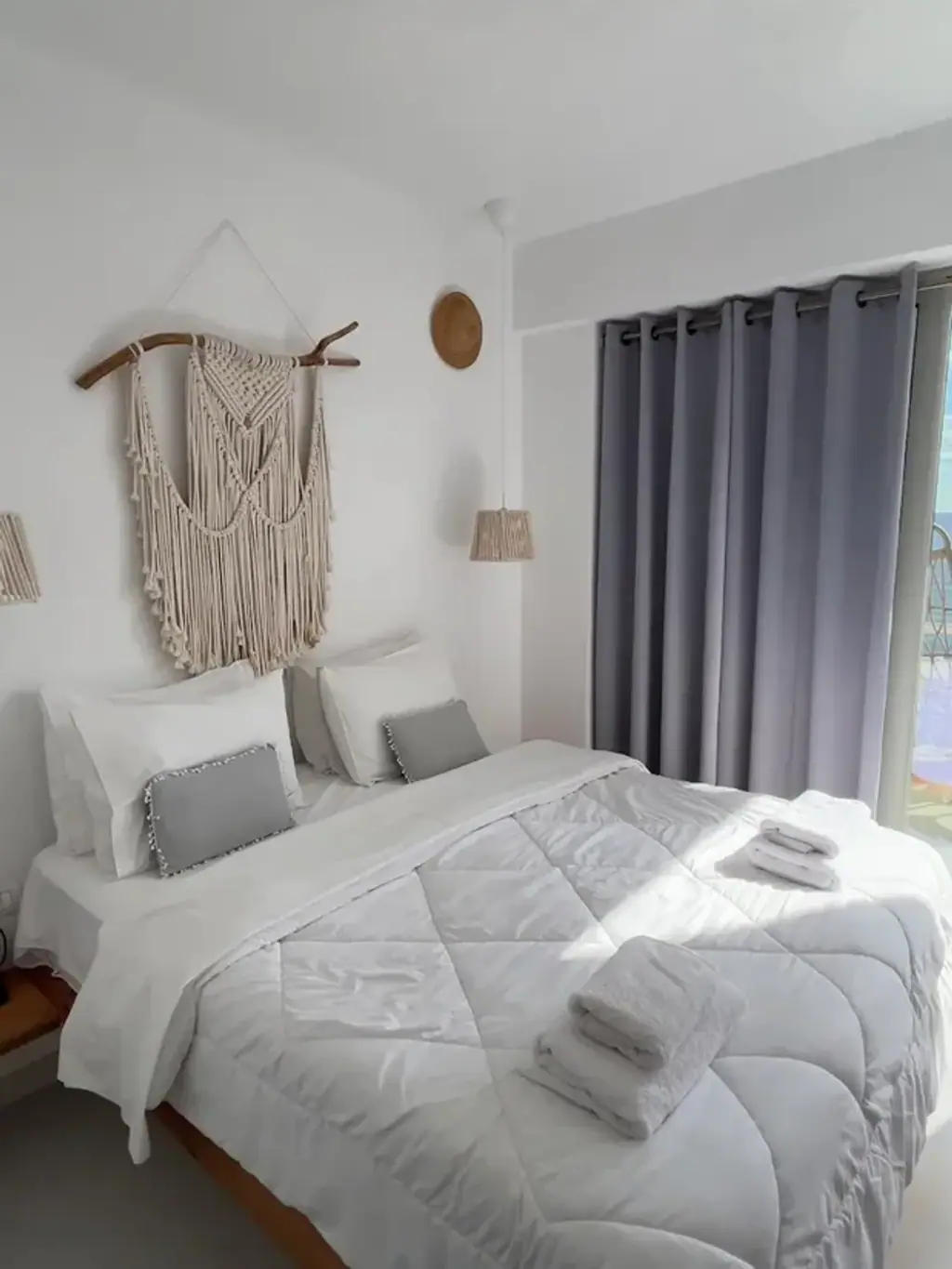 A hotel room with white walls, a macramé hanging over the bed and a grey curtain drawn across the balcony door