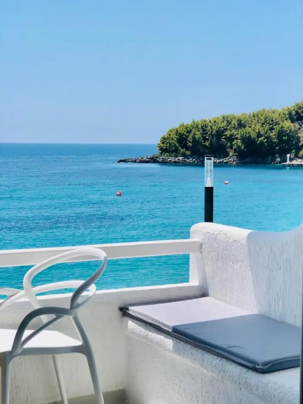 Turquoise water, a rock breakwater and a pine-covered headland seen over the white rail of a balcony