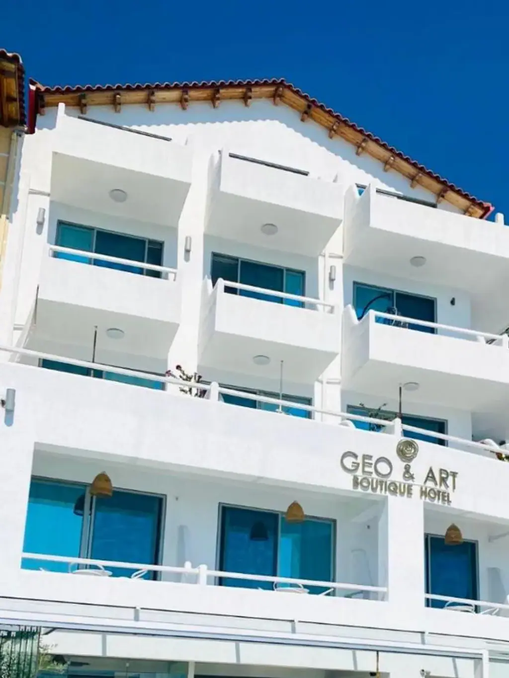 The white rendered front of Geo & Art Boutique Hotel, three tiers of balconies under timber eaves and terracotta pantiles, with the gilt GEO & ART sign