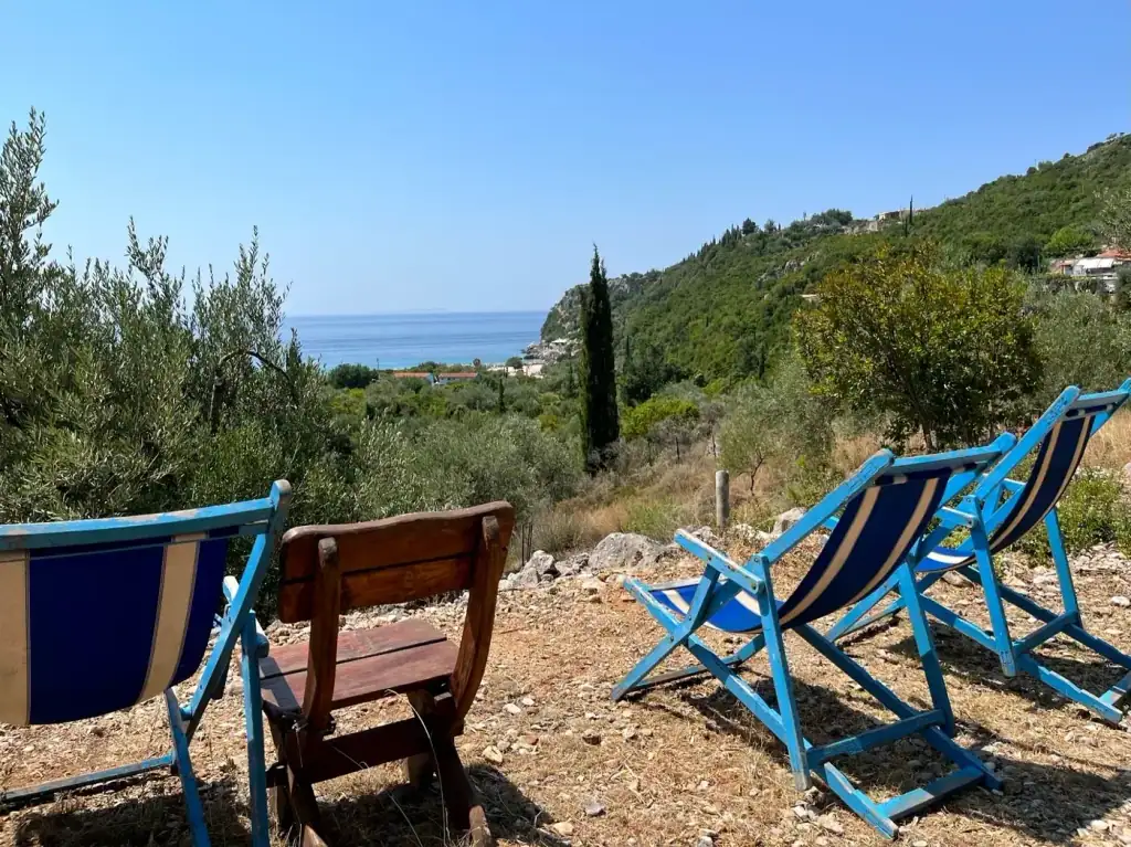 Striped blue deck chairs and a wooden bench on stony ground under olive trees, looking down a green valley to the bay at Livadhi