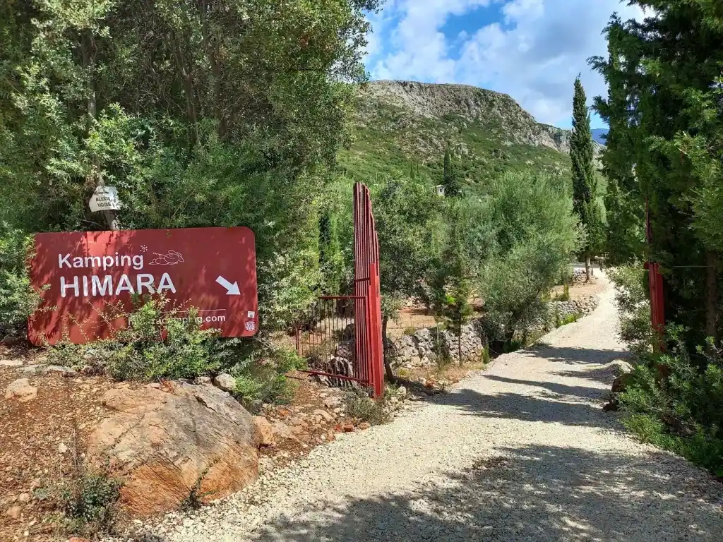 The dark red Kamping HIMARA sign board and red steel gate at the entrance, gravel drive climbing into the grove behind