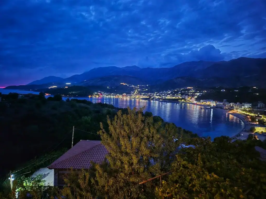 Himarë bay at dusk seen from the hillside, the town lights strung along the far shore and their reflection running out across the water