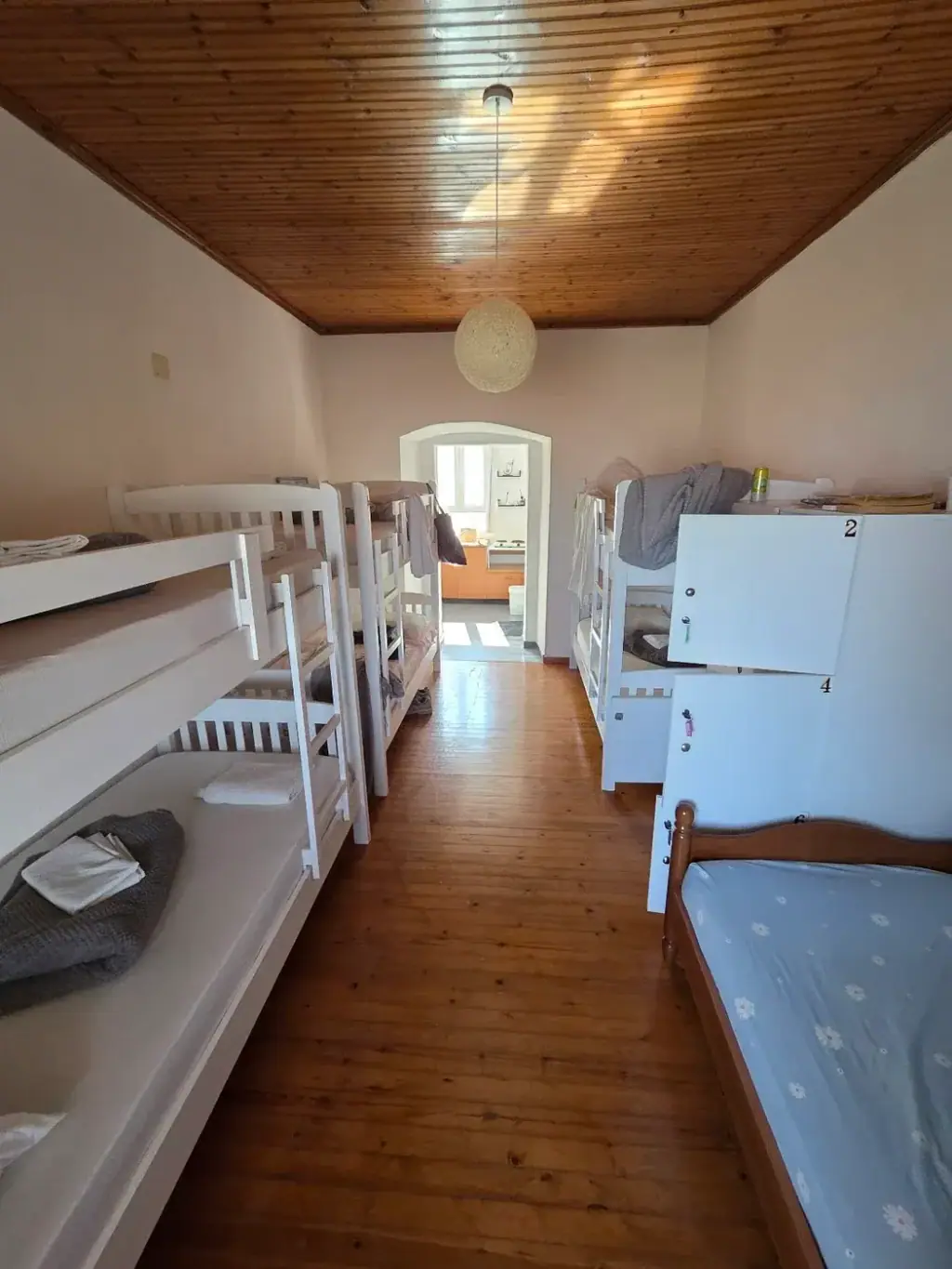 A dormitory with white bunk beds along both walls, a wooden ceiling, a wooden floor, numbered white lockers and an arched doorway at the far end
