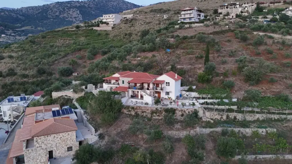 A white two-storey house with a red tiled roof and balconies, standing on a stone-walled terrace among olive trees on a dry hillside