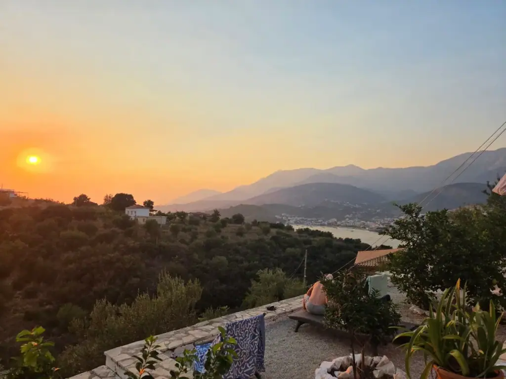 Sunset from a stone-walled terrace, the sun low over a ridge, hazy mountain ranges receding and a bay below with a town along its far edge