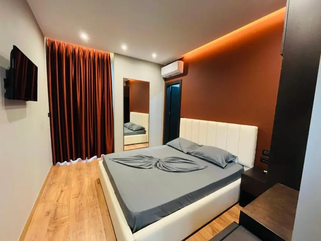 A room with grey bedding on a white padded headboard, a rust-red floor-length curtain, a wall-mounted television and a mirror