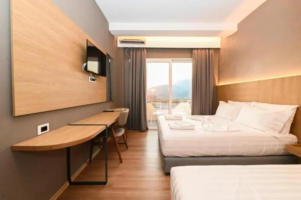 A refitted bedroom with an oak-panelled wall, a wall-mounted television, a long desk and a glazed balcony door showing hills and haze.