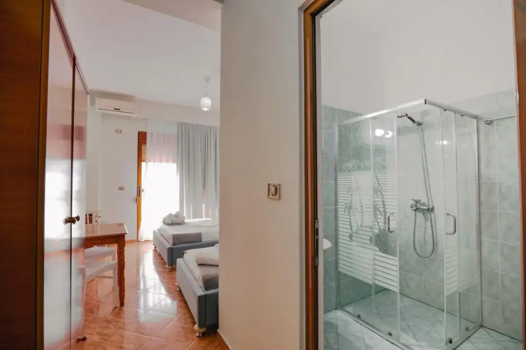 A corner shower cabin with etched sliding glass, seen through a doorway on the right; on the left a timber wardrobe with frosted panels, and between them the tiled bedroom with its curtained balcony door.