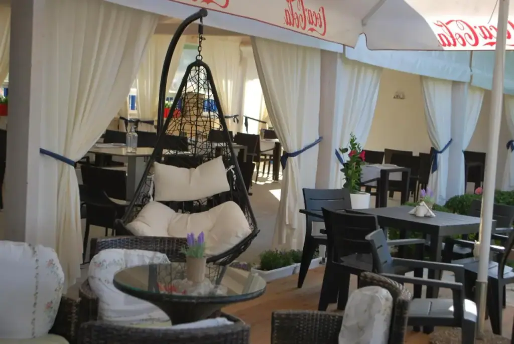 A hanging wicker chair beside dark tables and chairs in a room curtained in white