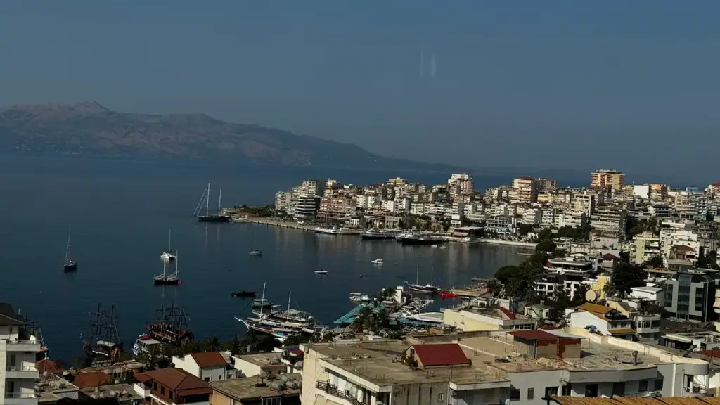 Saranda seen from above, apartment and hotel blocks packed down a steep hillside to the bay, with moored boats and the headland beyond