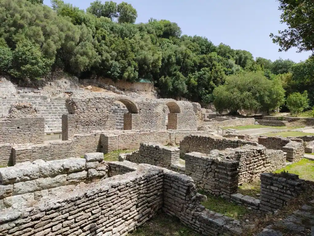 Brick arches of the Roman baths at Butrint with the stone seats of the ancient theatre behind them under thick green trees