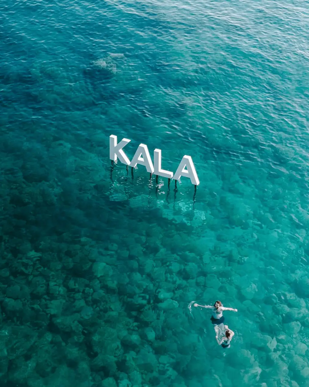 Aerial view of the iconic KALA letters standing in the turquoise shallows off Dhermi beach, with swimmers floating nearby, the Albanian Riviera setting of Kala Festival