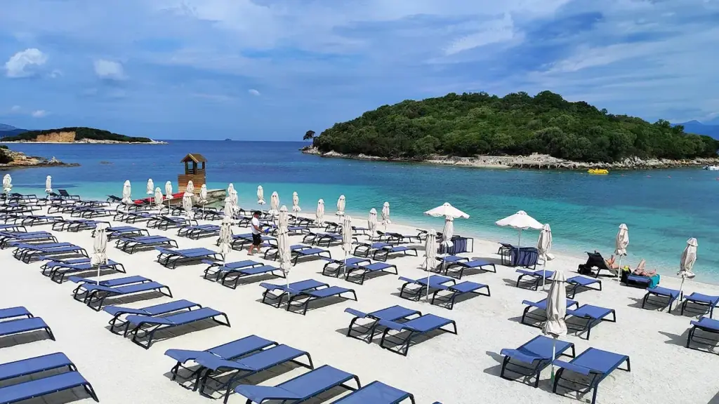 Neat rows of empty blue sun loungers and parasols on Ksamil's pale sand beside a small wooded islet and turquoise water