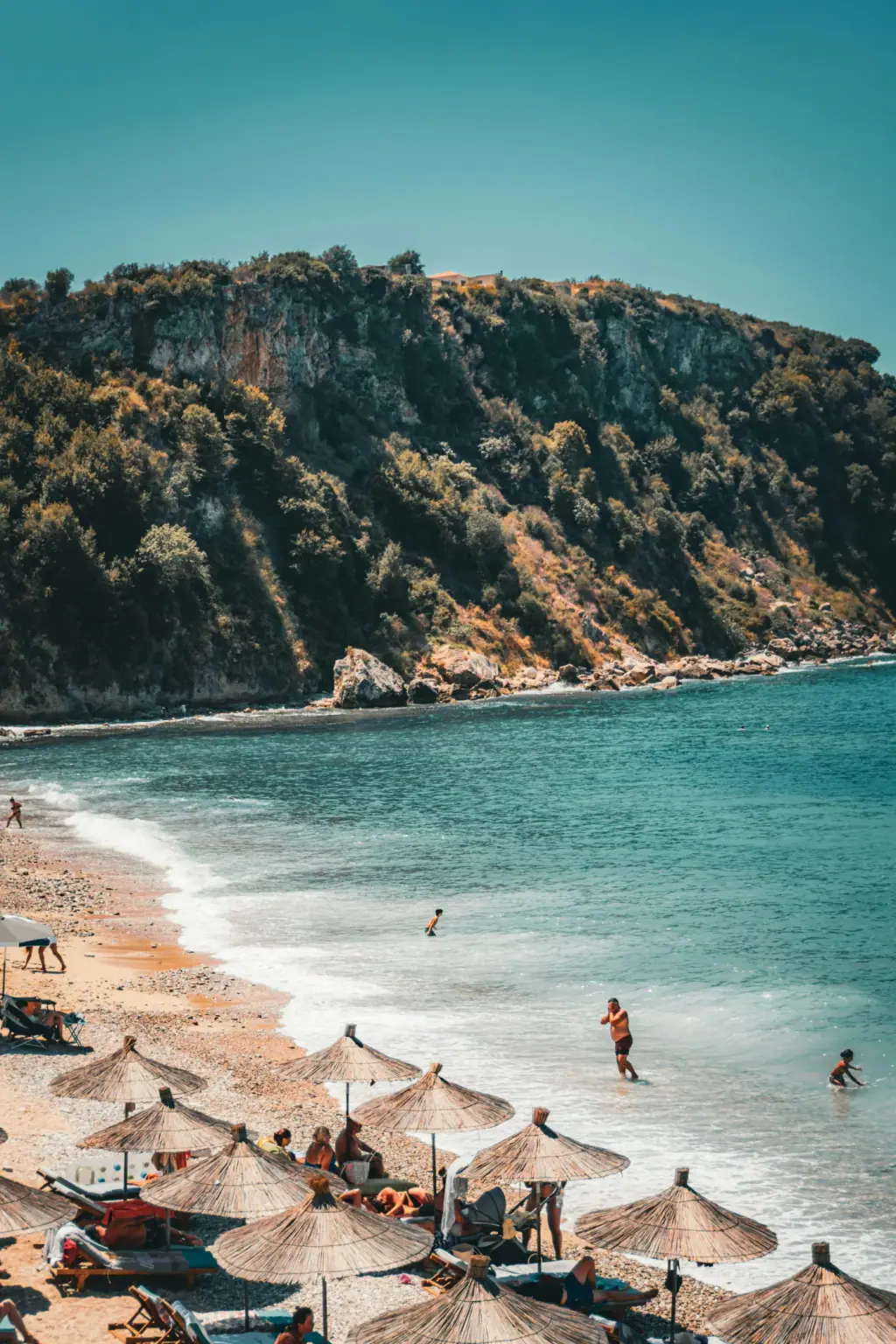 Thatched sun umbrellas and swimmers on a turquoise-water beach in southern Albania