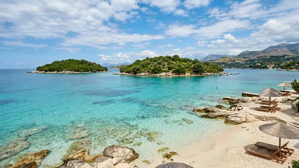 Clear turquoise shallows over pale rocks and sand, two small wooded islands offshore and thatched umbrellas on the beach under a cloud-streaked sky