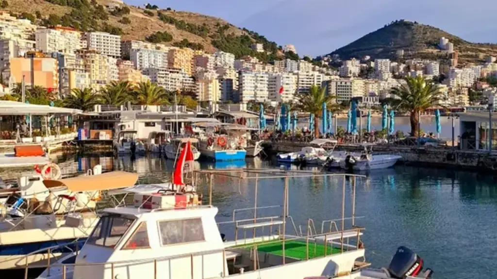 Saranda bay packed with boats and swimmers under a hard August sky, the promenade and hillside town rising behind