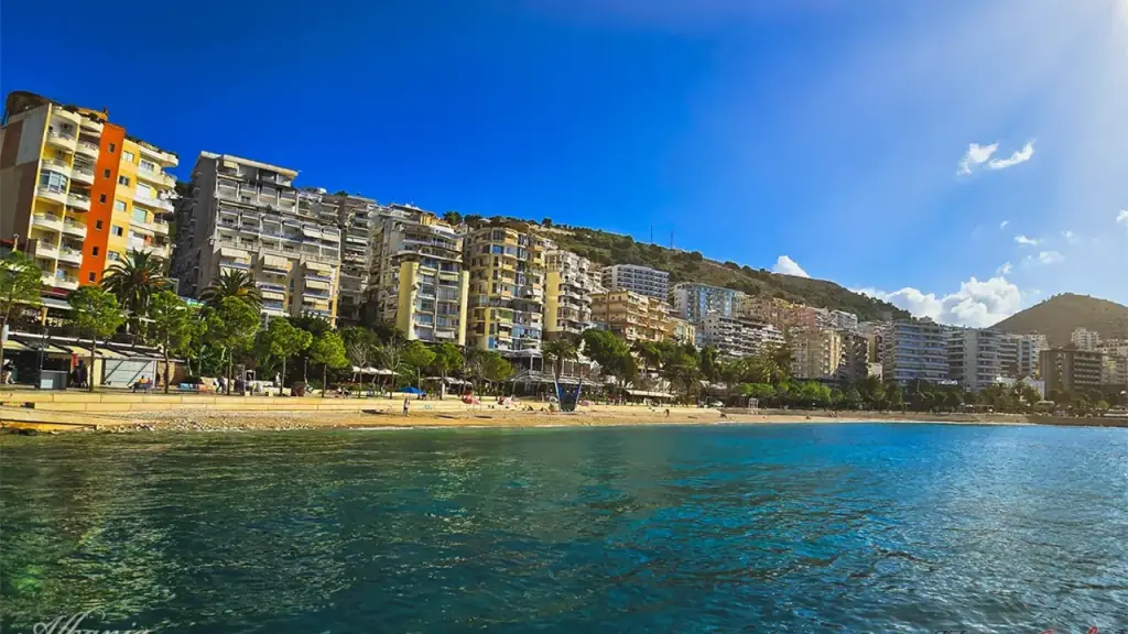 Saranda's curved bay and promenade in bright July light, hillside apartment blocks above calm blue water