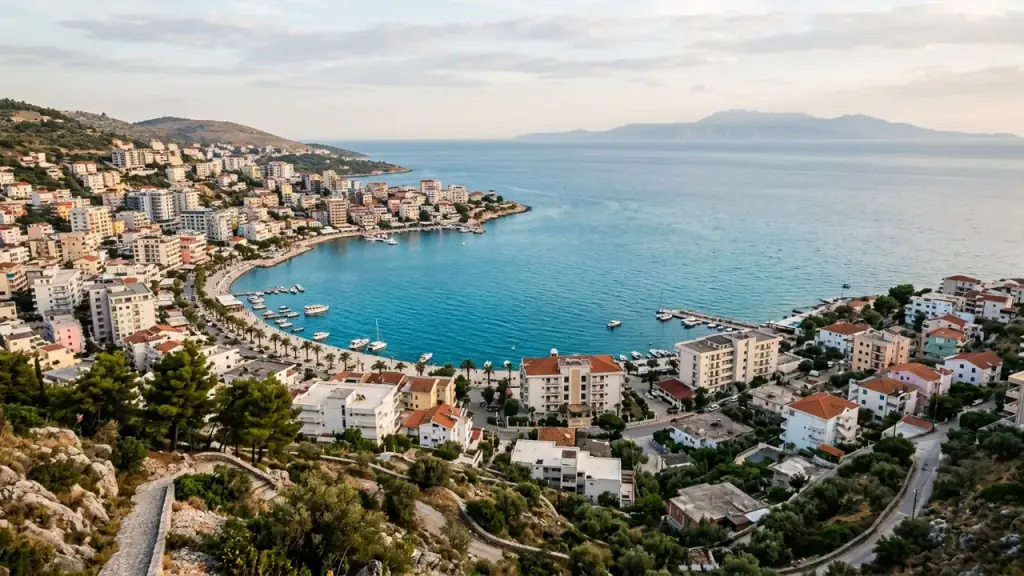 A curved bay lined with apartment blocks and a palm-lined promenade, calm turquoise water and a mountainous island on the horizon under evening cloud