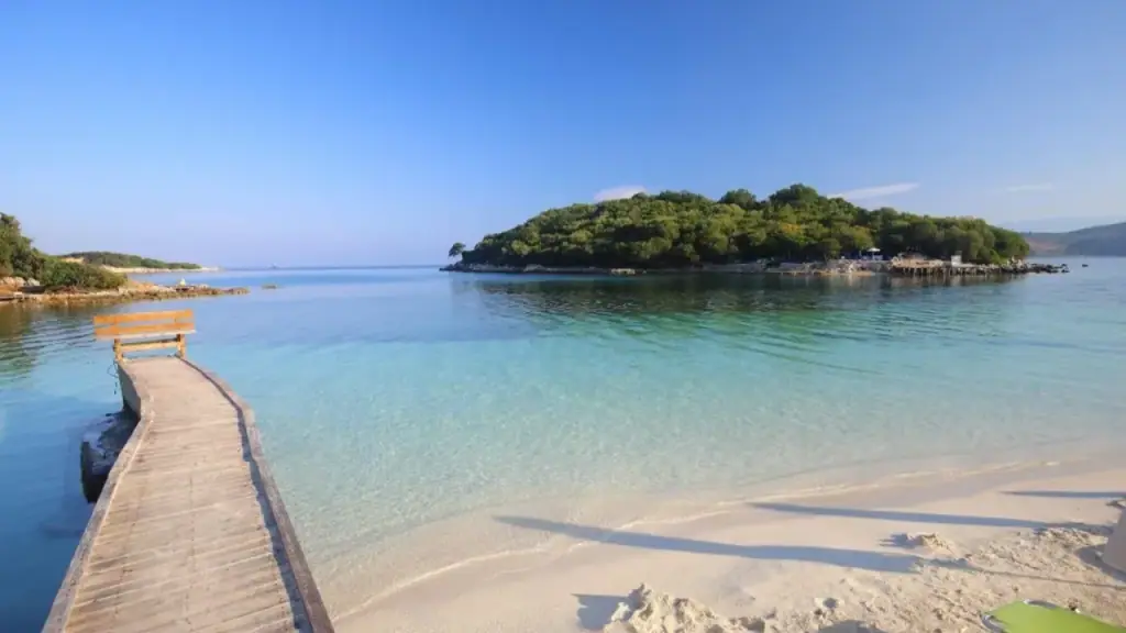 A short wooden jetty running out from Ksamil's pale sand towards a small wooded islet across clear shallow water