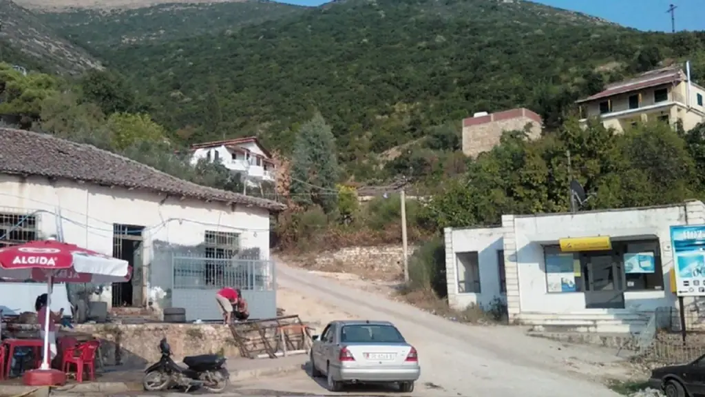 Small Riviera village square near Lukova with a car and motorbike parked on the dirt road — the kind of everyday transport Uber's app never shows in Albania