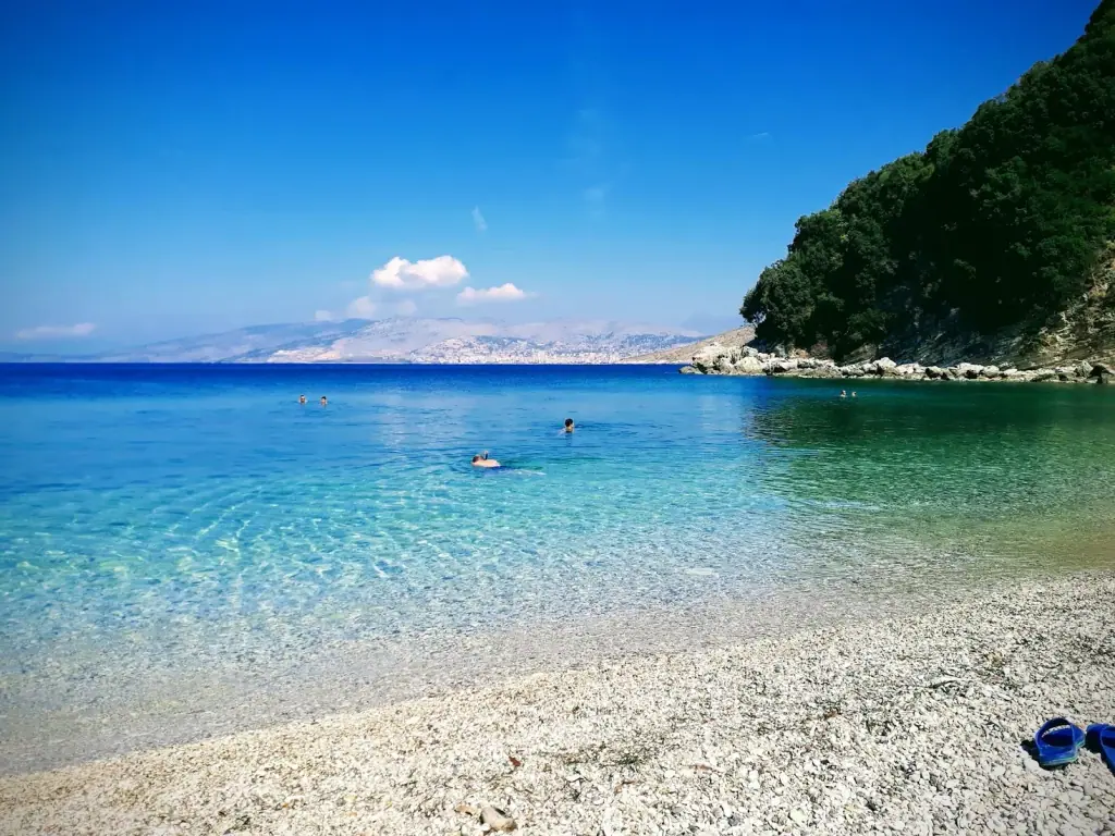 White pebble shore at Manastiri Beach with swimmers in clear shallow water and the hills across the bay