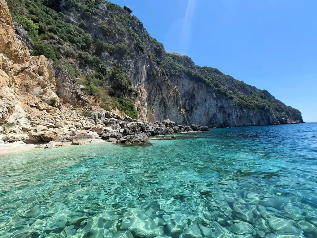 Clear shallow water over pale stones at Alevra Beach, with a high limestone cliff wall running out to sea behind it