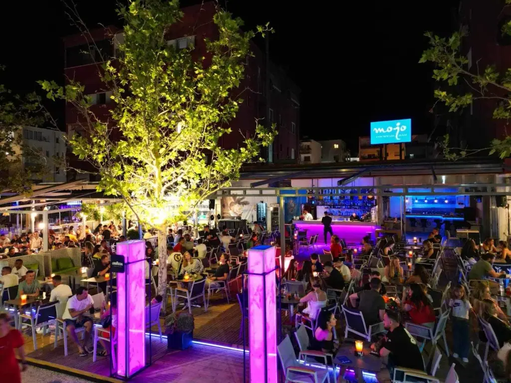 Busy evening terrace at Mojo Cocktails Bar with the illuminated Mojo sign and central bar in Spile