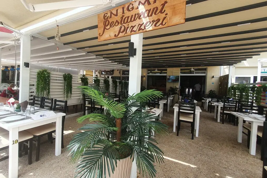 Covered terrace with the wooden Enigma Restaurant Pizzeria sign and set tables in Himara