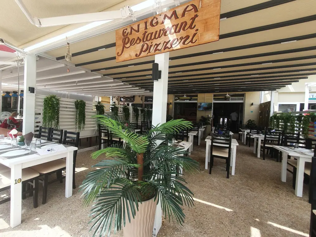 Covered terrace with the wooden Enigma Restaurant Pizzeria sign and set tables in Himara