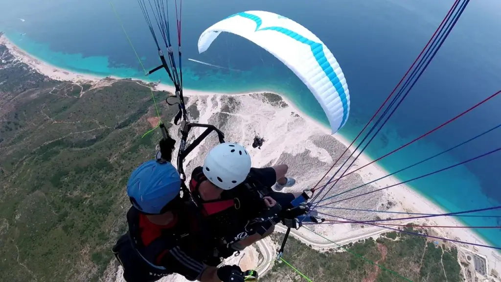 A tandem paraglider in flight above the white shingle and turquoise shallows of the Riviera coast