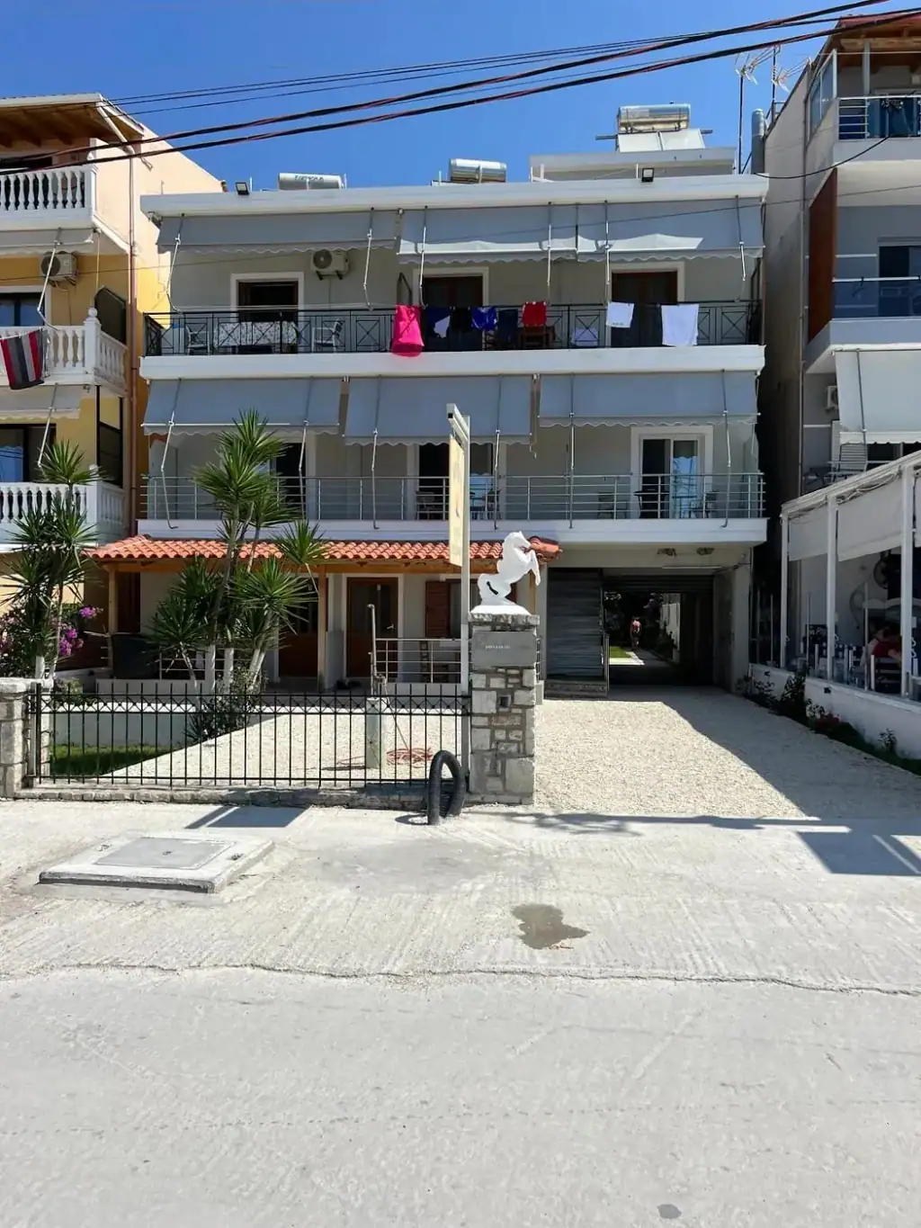 The front of the building from the beach road, awnings over every balcony and a gravel forecourt behind an iron railing